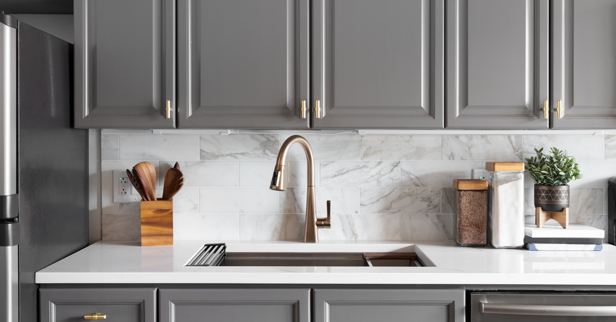 A kitchen features charcoal gray cabinets, white marble countertops, and a sink with a copper faucet.