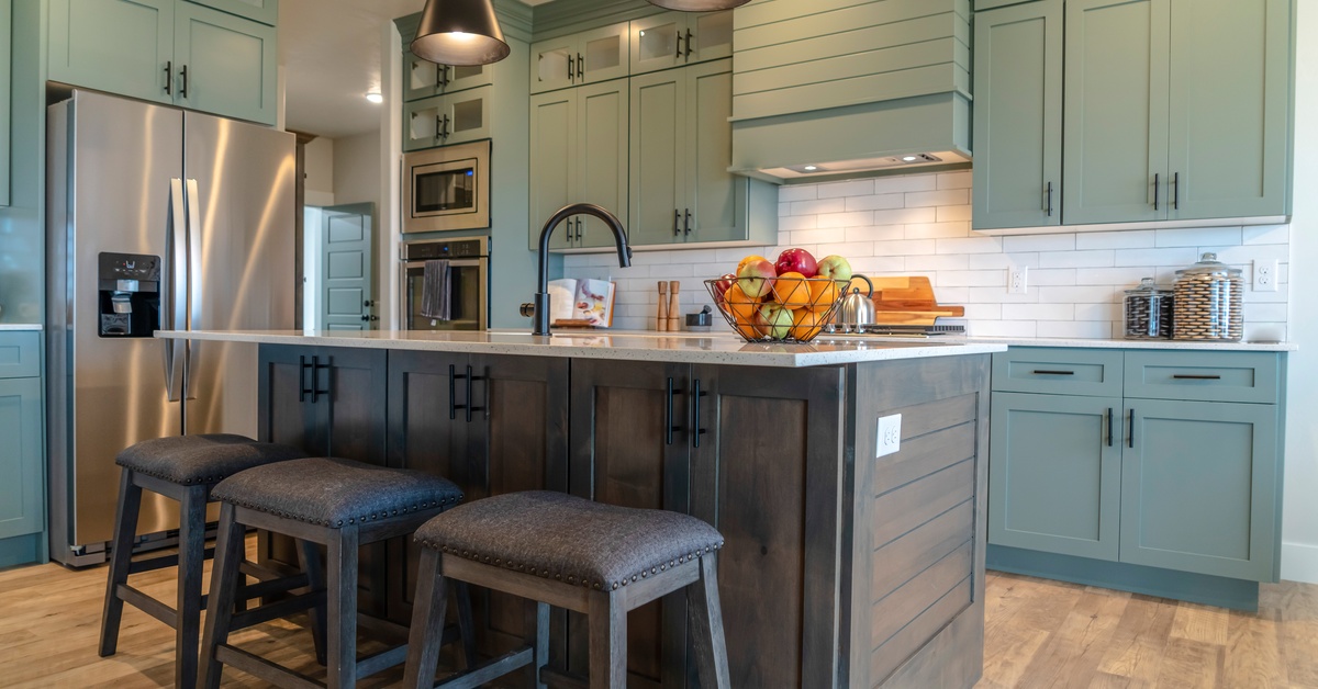 A cozy kitchen with teal upper cabinets, wooden lower cabinets, and dark stools and appliances that match the warm wood tones.